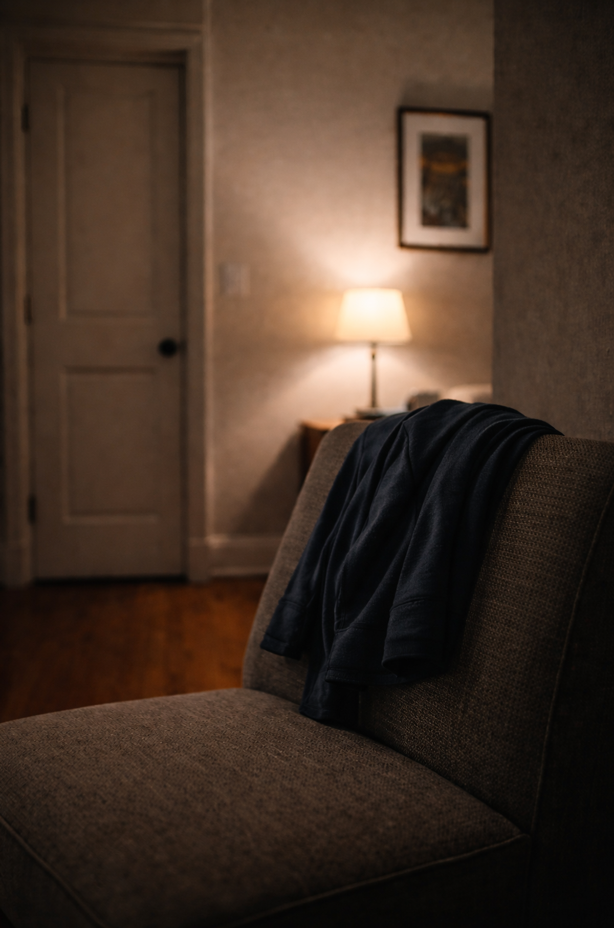 An empty armchair with a jacket draped over the back, softly lit by a nearby lamp in a quiet room, suggesting rest, absence, and unspoken interior weight.